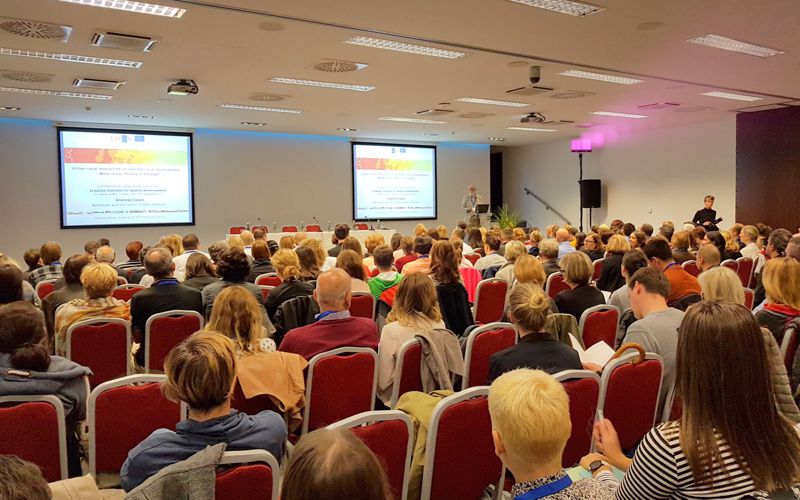 Large conference hall in cinema setup with rows of chairs, full audience and two presentation screens at the front