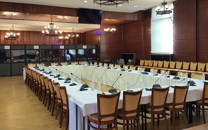 Conference hall with rectangular round table setup, fixed microphones at each seat, and interpreting booths visible in the background